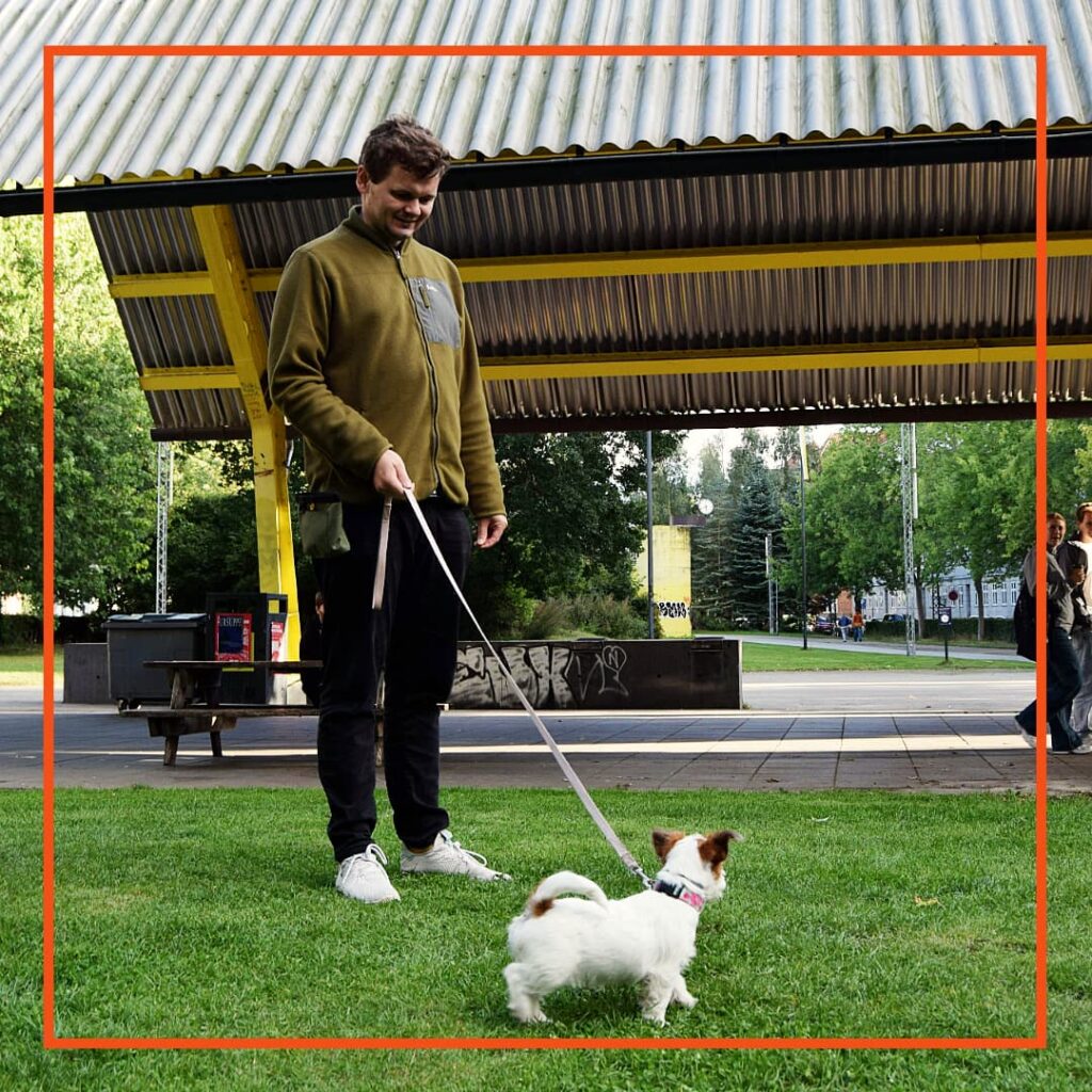 En deltager hos Klub Hund står med sin lille terrier i snor under hundetræning i parken. Hunden kigger opmærksomt på føreren, hvilket viser samarbejdet og kontakten, som er en central del af træningen hos Klub Hund.