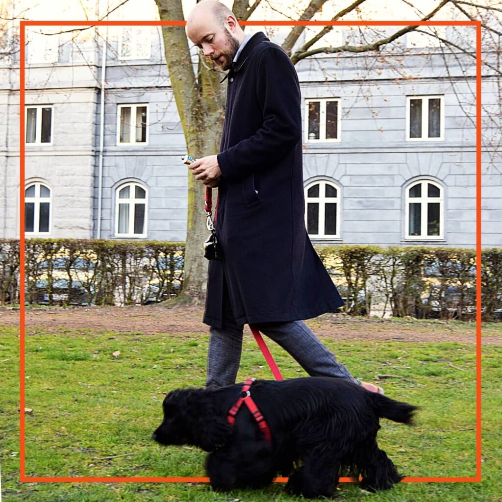 En deltager i hundetræning hos Klub Hund går en rolig tur med sin sorte hund i snor. Hunden går tæt ved ejerens side med fokus, mens ejeren smilende følger med og belønner god adfærd undervejs.
